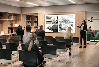 A woman presents slides on a screen to a small audience seated in a modern conference room with wooden flooring and bookshelves.