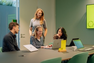 Four people collaborating around a conference table, using laptops, with one person standing and three seated, engaging in a discussion.