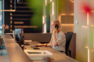 A woman at a modern reception desk is talking on the phone, surrounded by computers, with soft lighting and blurred background details.
