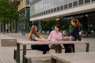 Three people sit and chat at an outdoor wooden picnic table, drinking and smiling, with a modern building in the background.