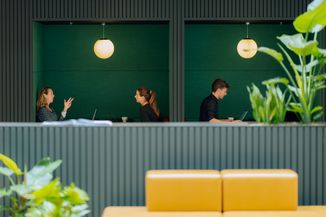 Two people converse in a modern green booth, while another works on a laptop nearby. Green plants frame the scene.