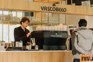 Barista making coffee at VascoBelo café counter, with coffee cups on the machine and a customer waiting.