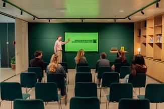 A person presents to a seated audience in a modern conference room with a green screen displaying "Offices have no future without people using them."
