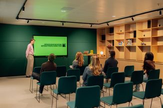 A person presents to an audience in a modern room with green decor. The screen reads, "Offices have no future without people using them."