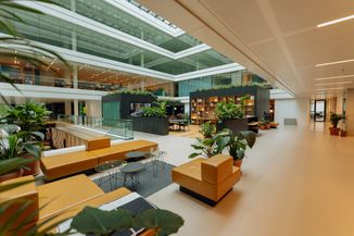 Modern office atrium with yellow seating, glass railings, and abundant greenery. Open workspace visible above through glass walls.
