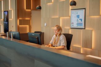 A woman is seated at a modern reception desk with wood paneling and ambient lighting, working on a computer.