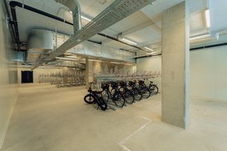 Spacious indoor bike parking area with several parked bikes and numerous empty metal racks, under bright industrial lighting.