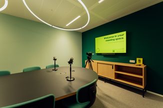 Modern meeting room with oval table, microphones, green chairs, and a digital display reading "Welcome in your meeting space."