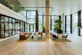 Modern lobby with brown sofas, chairs, and small tables on green rugs. Large windows, glass walls, and indoor plants enhance the open space.