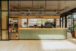 Modern coffee shop interior with a green-tiled counter, espresso machine, a fridge with drinks, and large windows in the background.