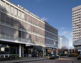 Modern office building "Berghaus Plaza" with large windows, bicycles, and parked cars on a sunny day, set against a clear blue sky.