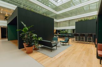 Modern indoor seating area with dark furniture, plants, and wood flooring, surrounded by glass walls in an open office setting.