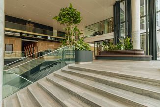 Spacious modern lobby with wooden floors, glass railings, large potted plants, and seating areas surrounded by glass-walled offices.