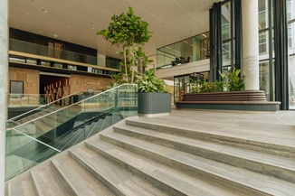 Spacious modern lobby with wooden floors, glass railings, large potted plants, and seating areas surrounded by glass-walled offices.