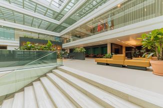 Modern atrium with glass walls, indoor plants, orange seating, and a staircase; flags displayed inside the spacious, well-lit area.