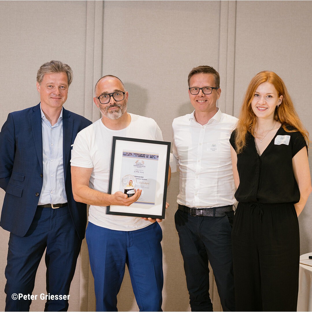 Quatro pessoas juntas, sorrindo e posando para uma foto. O homem no meio está segurando um certificado emoldurado.