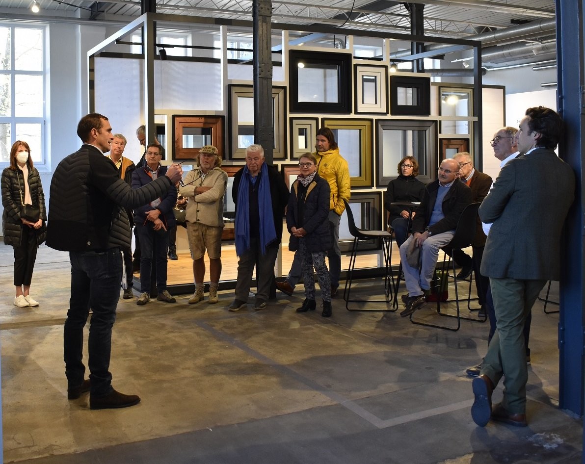 Eine Gruppe von Menschen hat sich in einer Kunstgalerie versammelt. Einige stehen, andere sitzen. Ein Mann in der Mitte spricht zur Gruppe. Die Galerie präsentiert eine große Installation von gerahmten Kunstwerken an der Wand. Der Boden ist aus Beton, und im Hintergrund stehen Stühle und ein Tisch.