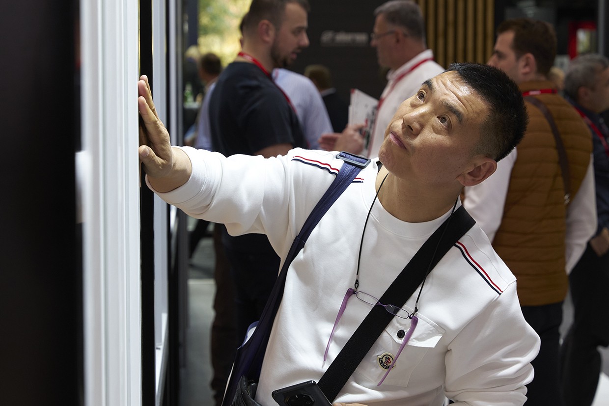 A man with a white shirt and a black strap around his neck is touching a white wall with his hand. He is looking up and seems to be checking something. Behind him, there are several people standing and looking at something. One of them is holding a piece of paper. They are probably in a conference room.