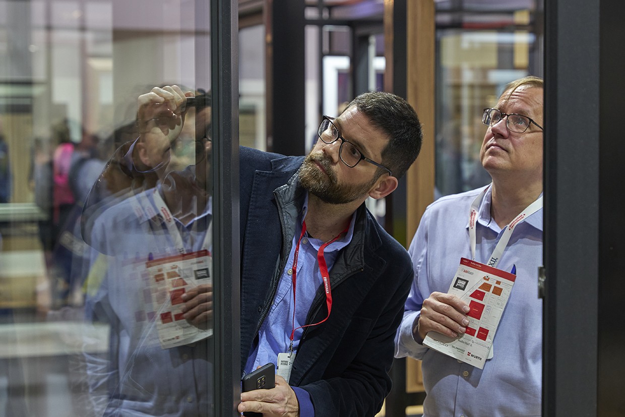 Dois homens com crachás em volta do pescoço estão parados em frente a uma porta de vidro, olhando para algo do outro lado.