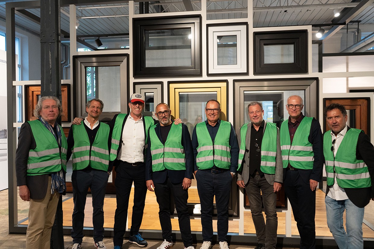 Seven men in green safety vests stand together in front of a wall of windows, smiling for the camera.