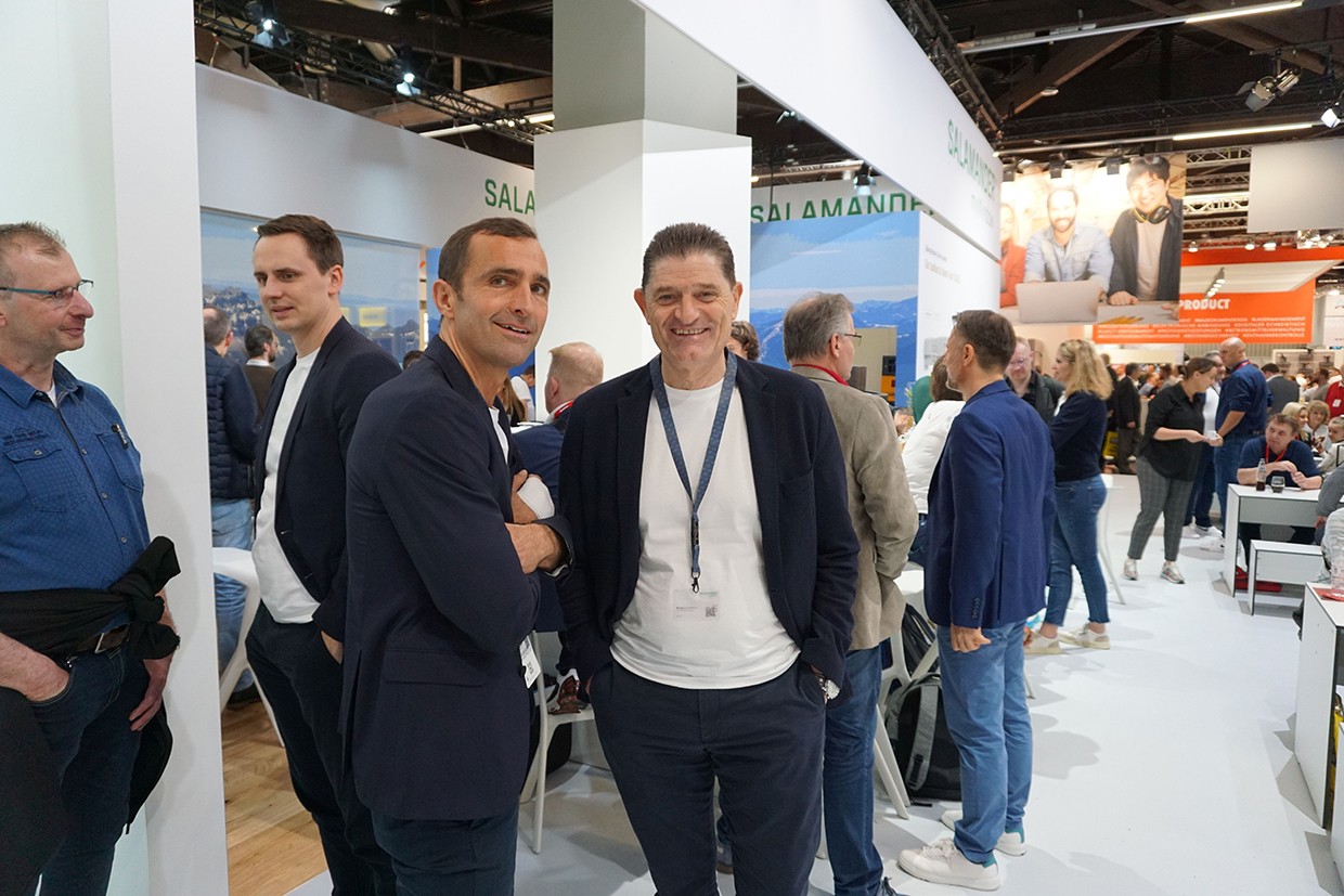 A group of men are standing together in a room. Two of them are smiling and posing for a photo.