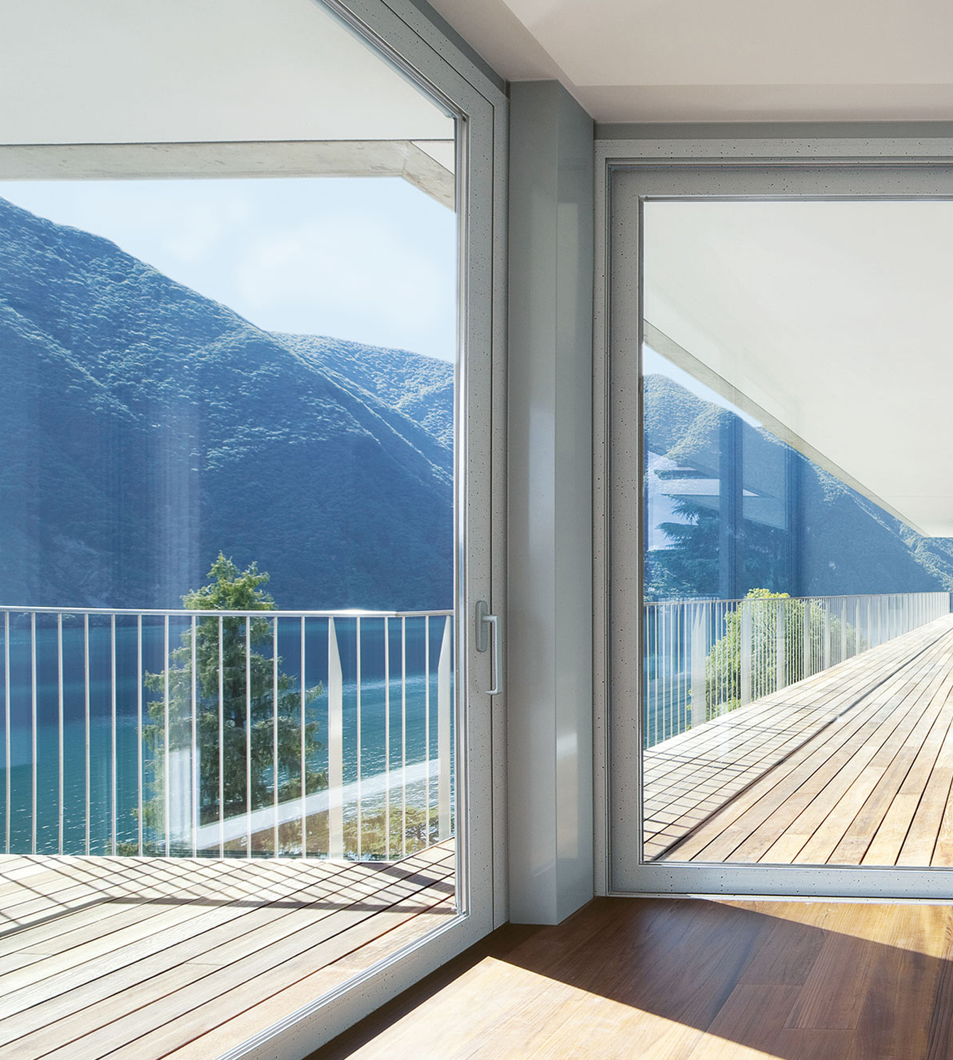 Portas de vidro deslizantes que levam a um deck de madeira com um corrimão de metal, com vista para um lago e montanhas.