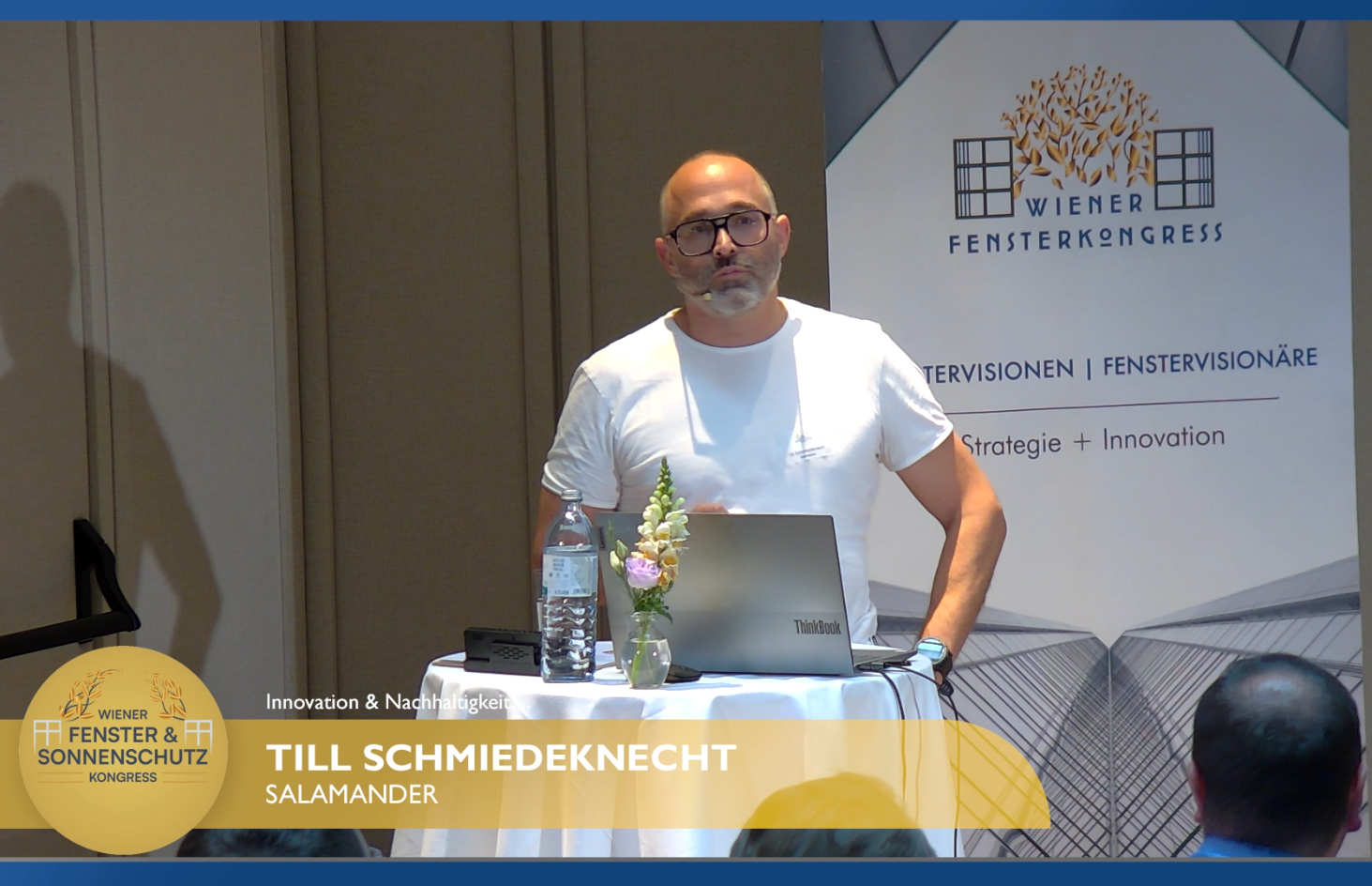 Um homem está atrás de uma mesa com um laptop, uma garrafa de água e flores, falando em um microfone em uma conferência.