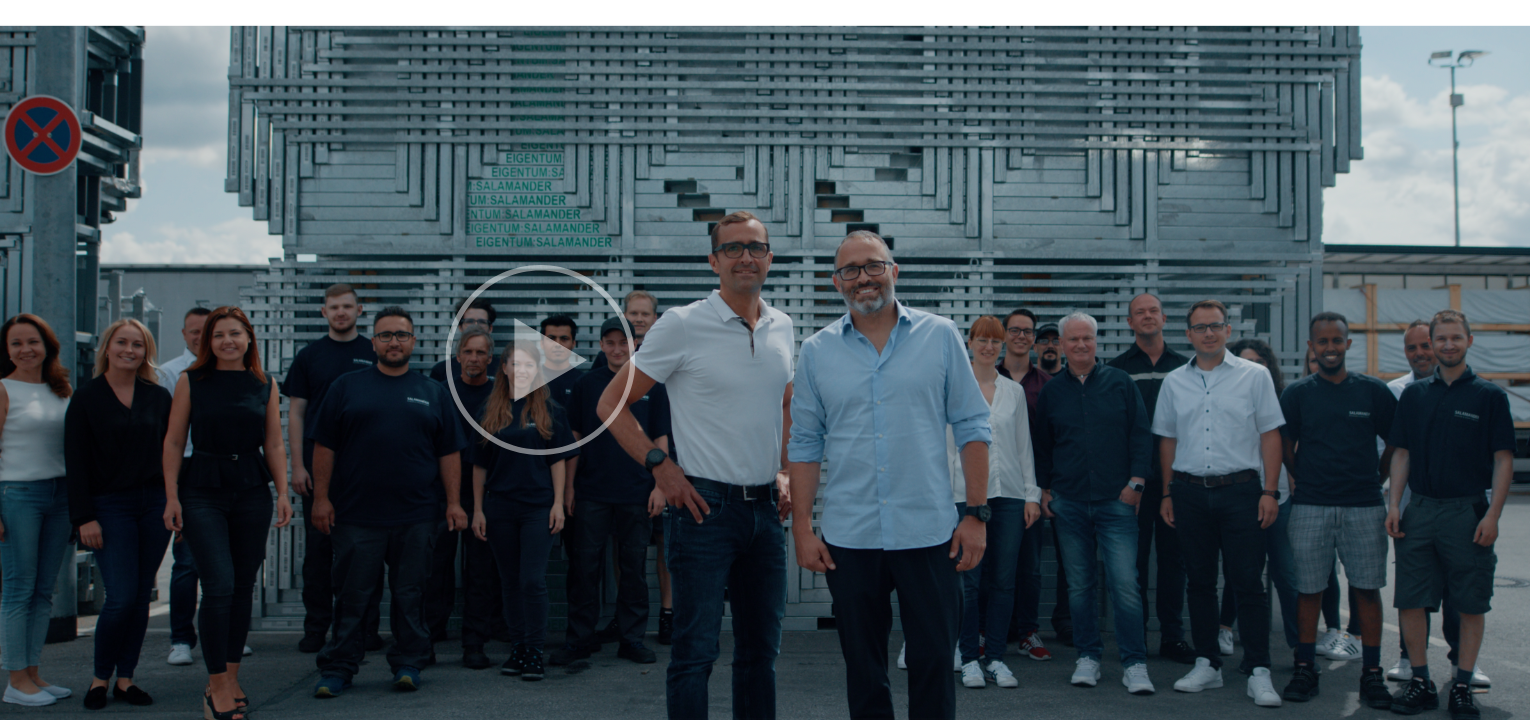 A group of people are standing in front of a building. Two men are standing in front of the group. The man on the right is wearing a blue shirt and black pants. The man on the left is wearing a white shirt and black pants. The group is wearing black shirts and pants. The building has a metal structure with a sign on top.