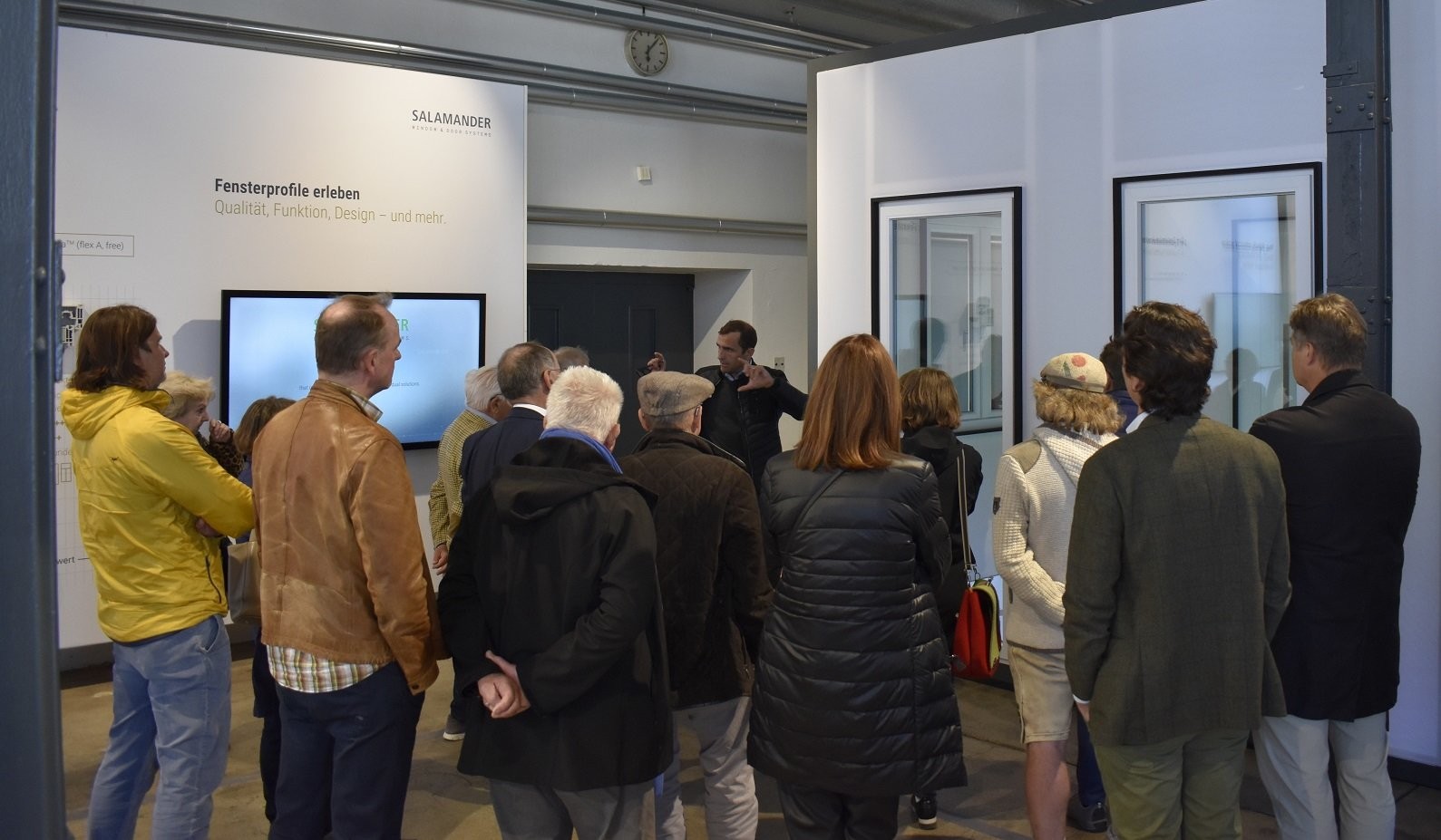 Um grupo de pessoas está reunido em uma sala, ouvindo um homem falar. O homem está em pé na frente de uma tela grande, e há duas fotos emolduradas na parede atrás dele. A sala tem uma parede branca e uma porta preta.