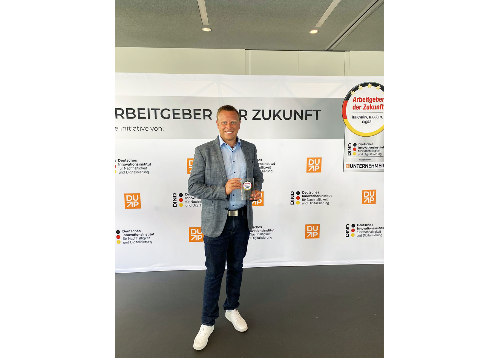 A man stands in front of a banner holding an award. The banner reads "Arbeitgeber der Zukunft" and "RBEITEBER R ZUKUNFT".