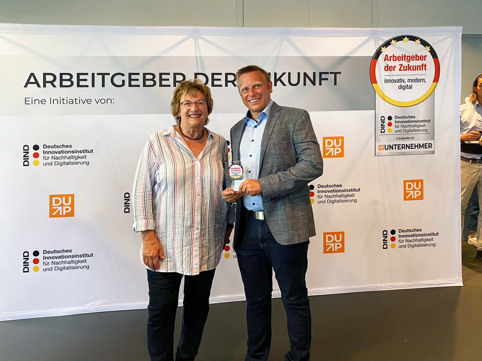 A man and a woman standing next to each other holding an award in front of a banner.
