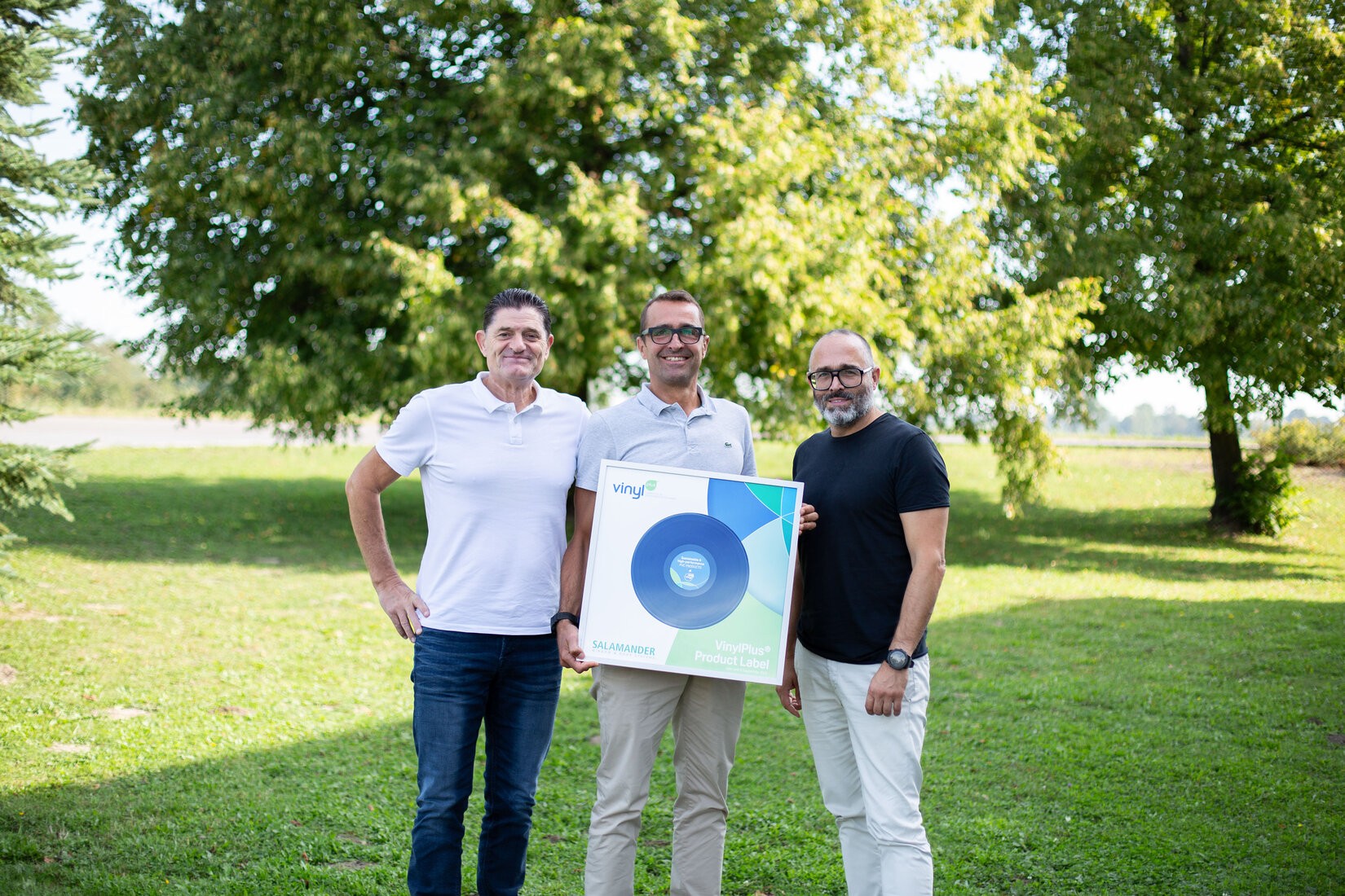 Three men standing in a park with trees in the background. The man in the middle is holding a framed vinyl record with the words "VinylPlus Product Label" written on it. The man on the left is wearing a white polo shirt, while the man on the right is wearing a black shirt and glasses.
