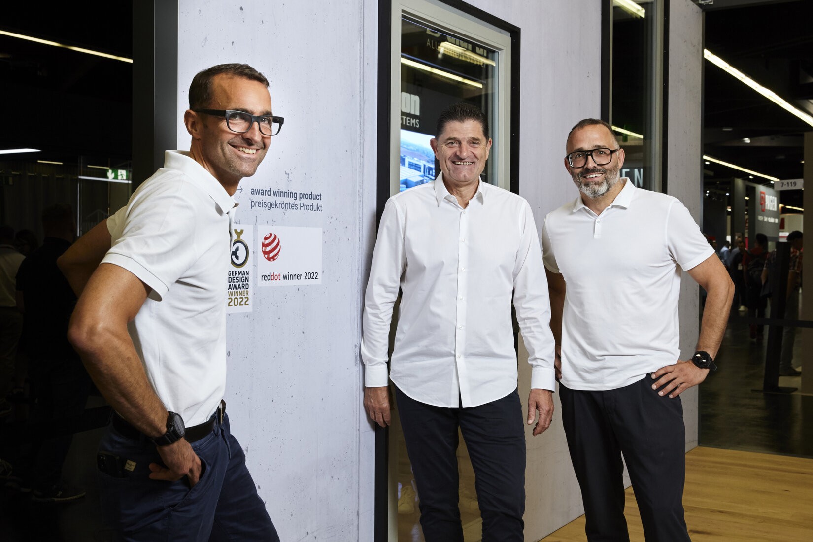 Three men in white shirts and black pants stand next to each other in front of a wall. The man on the left wears glasses and a watch, the man in the middle wears a watch, and the man on the right wears glasses and a watch. Behind them is a glass door with a sign that reads "All Systems On".