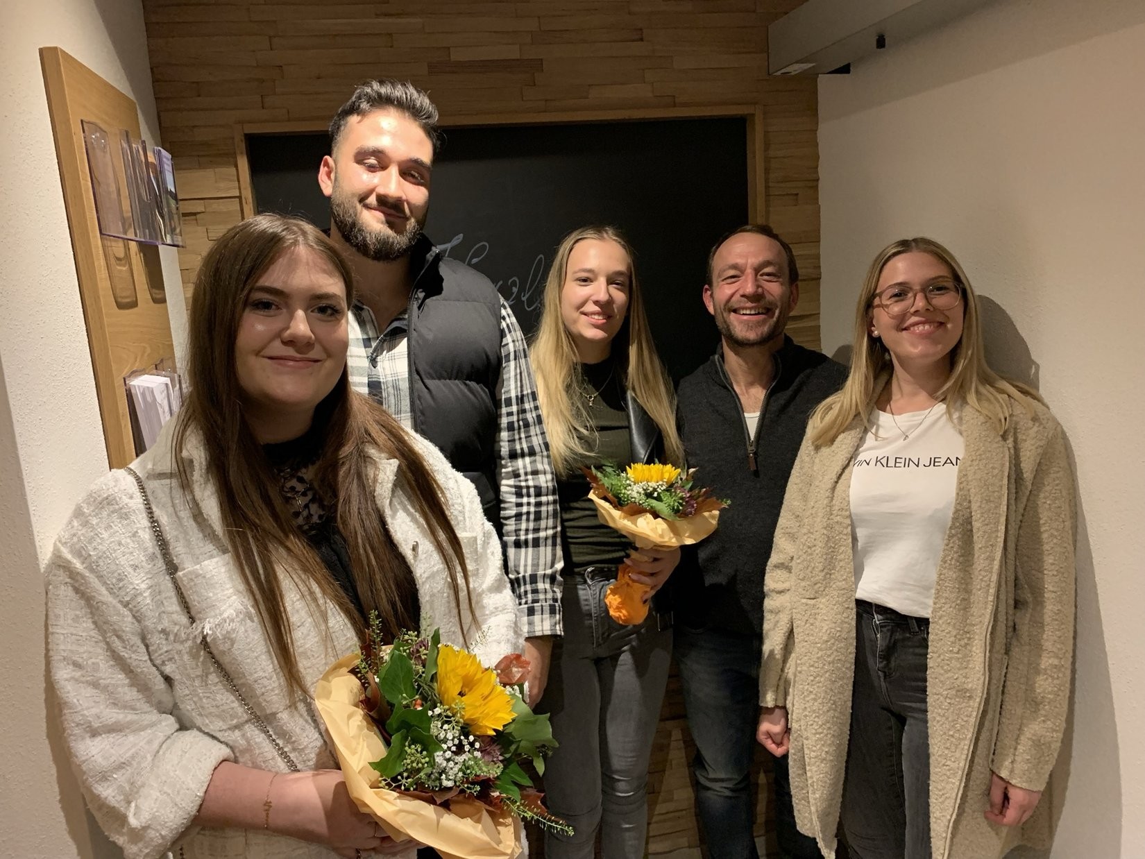 Five people standing together in a room, two of them holding flower bouquets, smiling and posing for a photo.