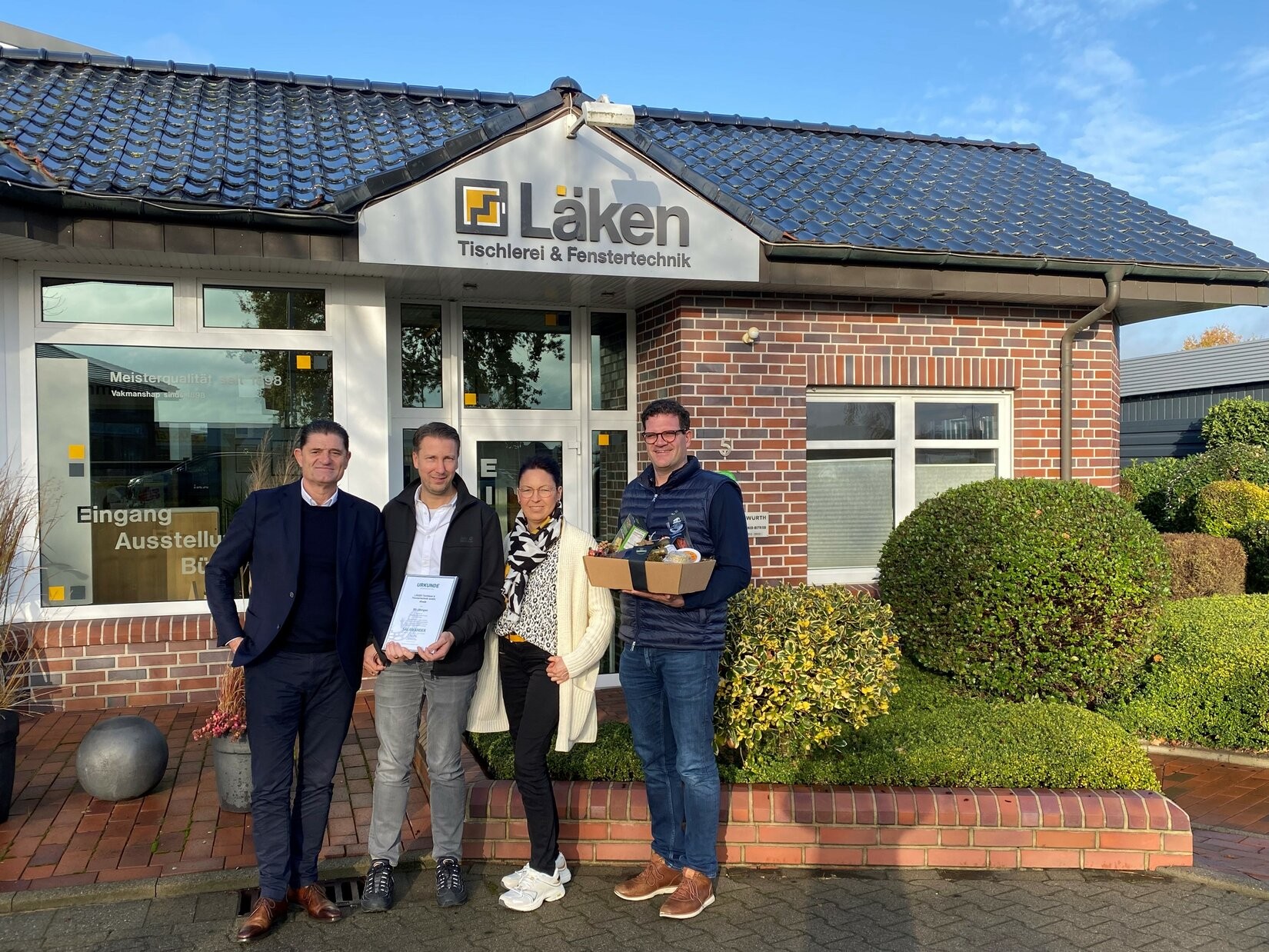 Quatro pessoas estão em pé em frente a um edifício com o nome Laken nele. Elas estão sorrindo e posando para uma foto.