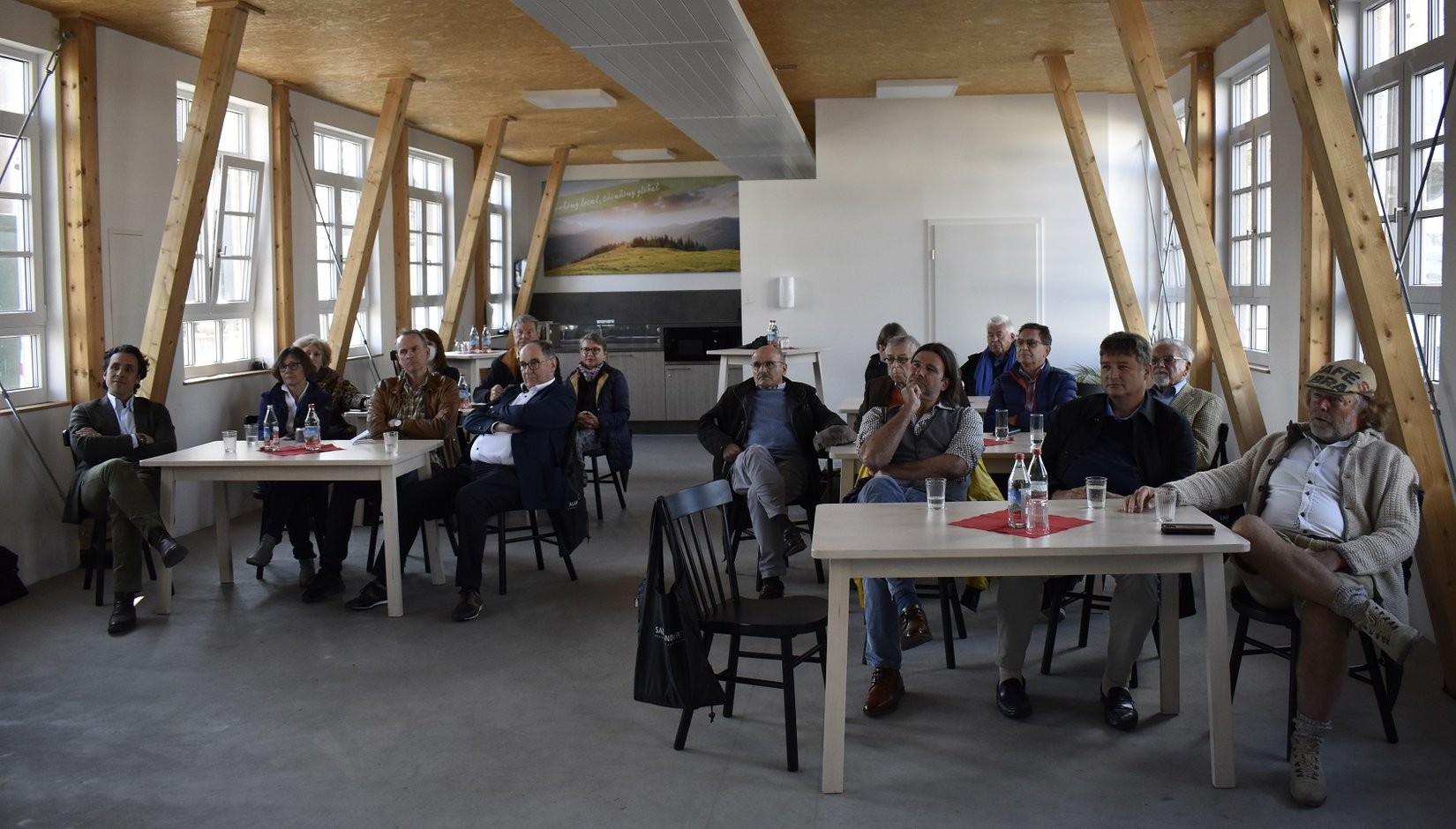 Eine Gruppe von Menschen sitzt an Tischen in einem Raum mit großen Fenstern und Holzbalken.
