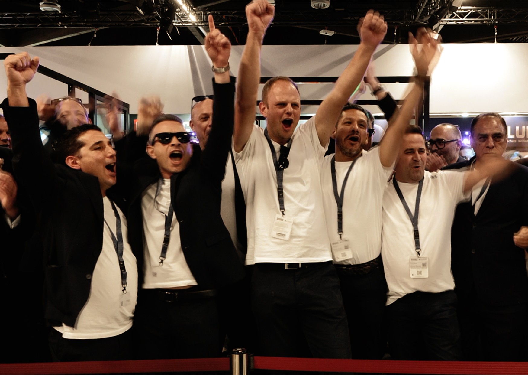 A group of men in formal attire are standing together, raising their hands in celebration. They are wearing white shirts and black pants, with some wearing sunglasses and others wearing ties. They are all wearing name tags around their necks. Behind them, there is a red rope barrier, and in the background, there are people standing and sitting, some of whom are wearing glasses.