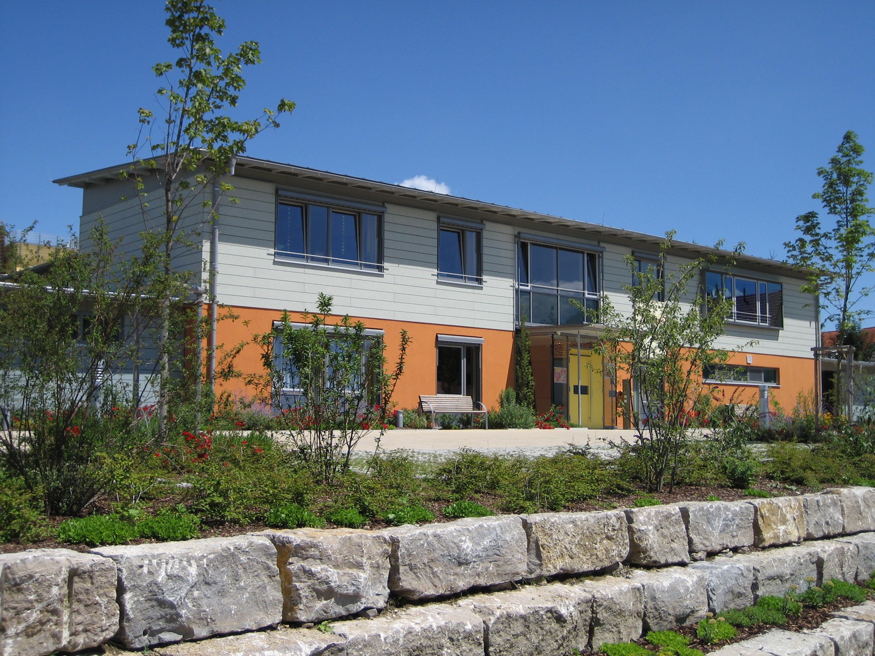 A modern building with a stone wall and a garden in front. The building has a yellow door and a bench in front of it. There are trees and plants in front of the building.