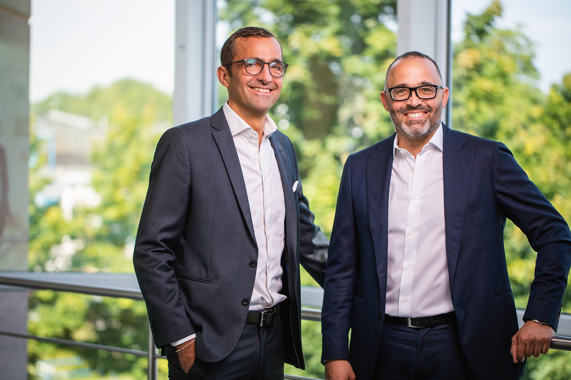 Dois homens em trajes de negócios estão de pé um ao lado do outro em frente a uma parede de vidro com árvores ao fundo.