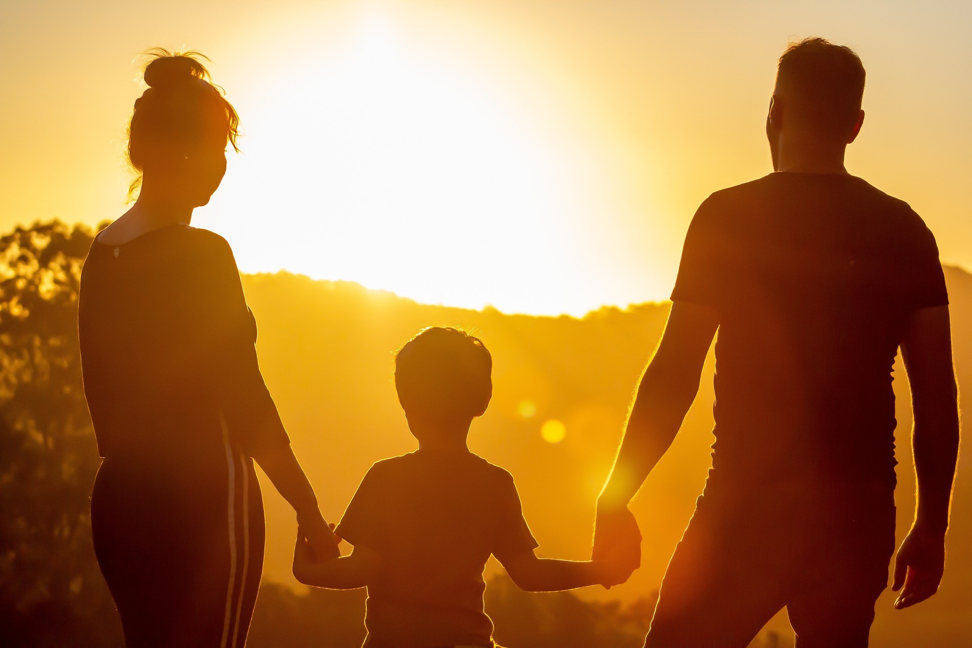 Silhouette of a family of three, consisting of a man, a woman, and a young boy, holding hands against a backdrop of a setting sun.