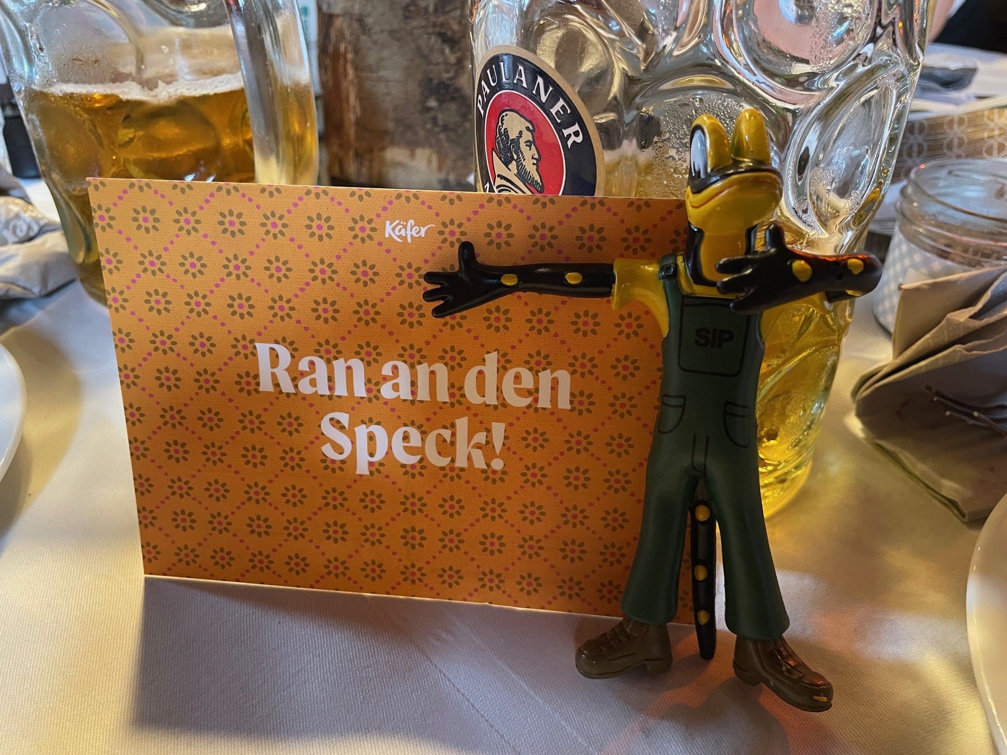 A figurine of a frog in overalls stands next to a sign that reads "Ran an den Speck!" in German.