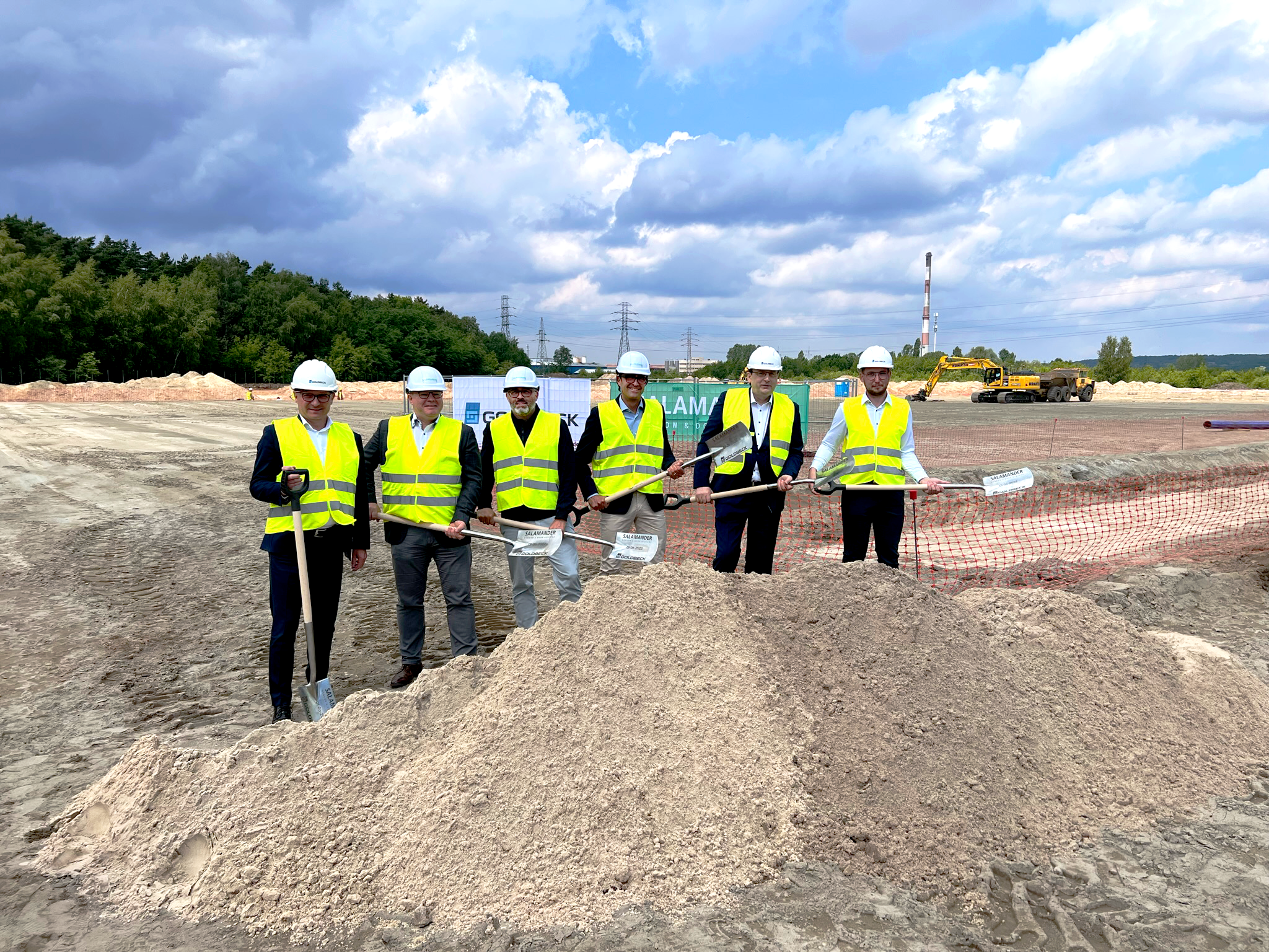 Sei uomini in giubbotti gialli e caschi bianchi stanno davanti a una pila di terra, tenendo pale.