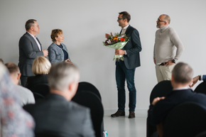 A man in a suit holding a bouquet of flowers stands in front of a group of people seated in chairs.