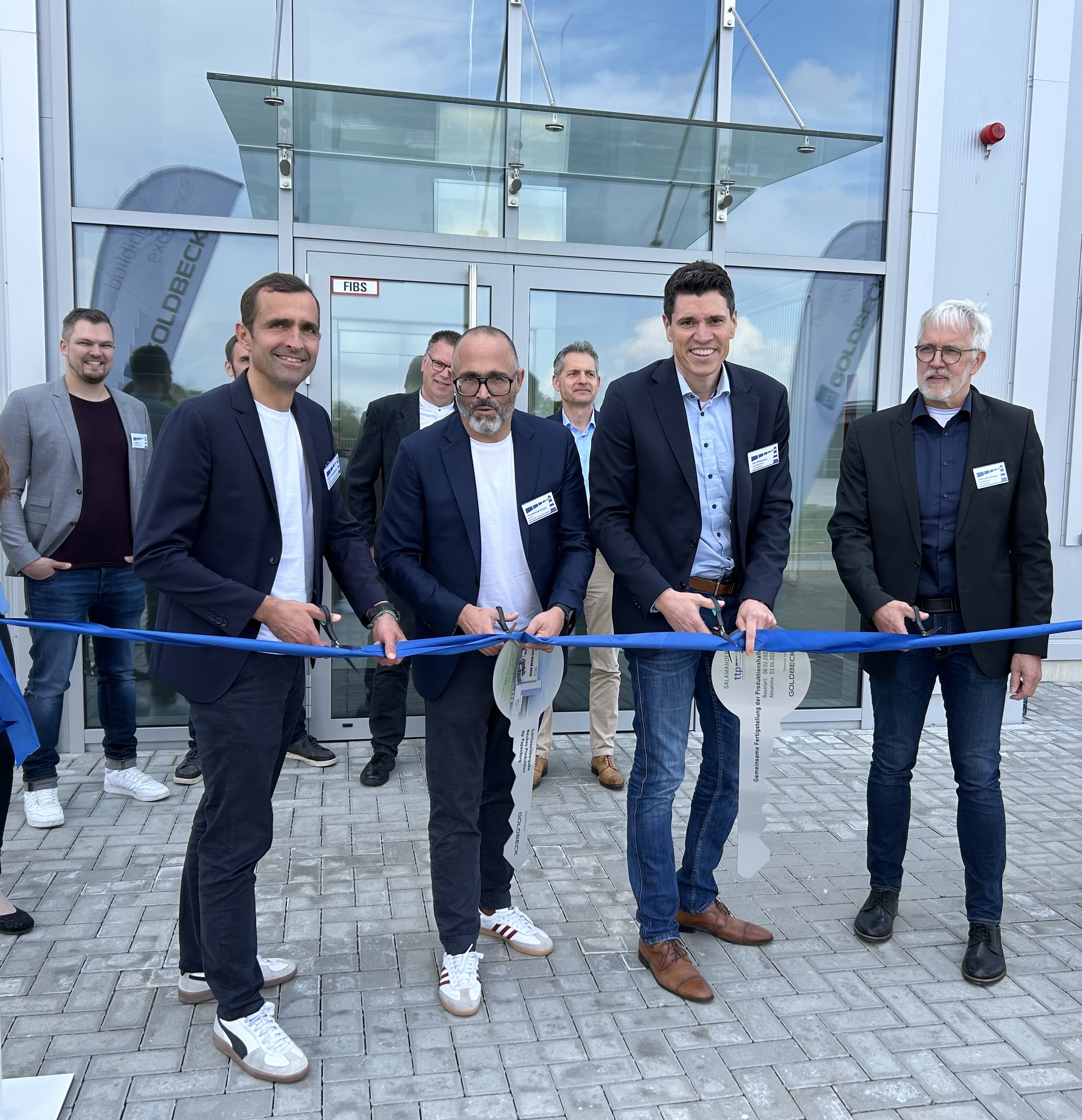 Quatro homens cortando uma fita em frente a um edifício com um grupo de pessoas atrás deles.