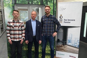 Three men standing next to each other in front of a banner. The man on the left is wearing a plaid shirt, the man in the middle is wearing a suit, and the man on the right is wearing a plaid shirt and jeans.