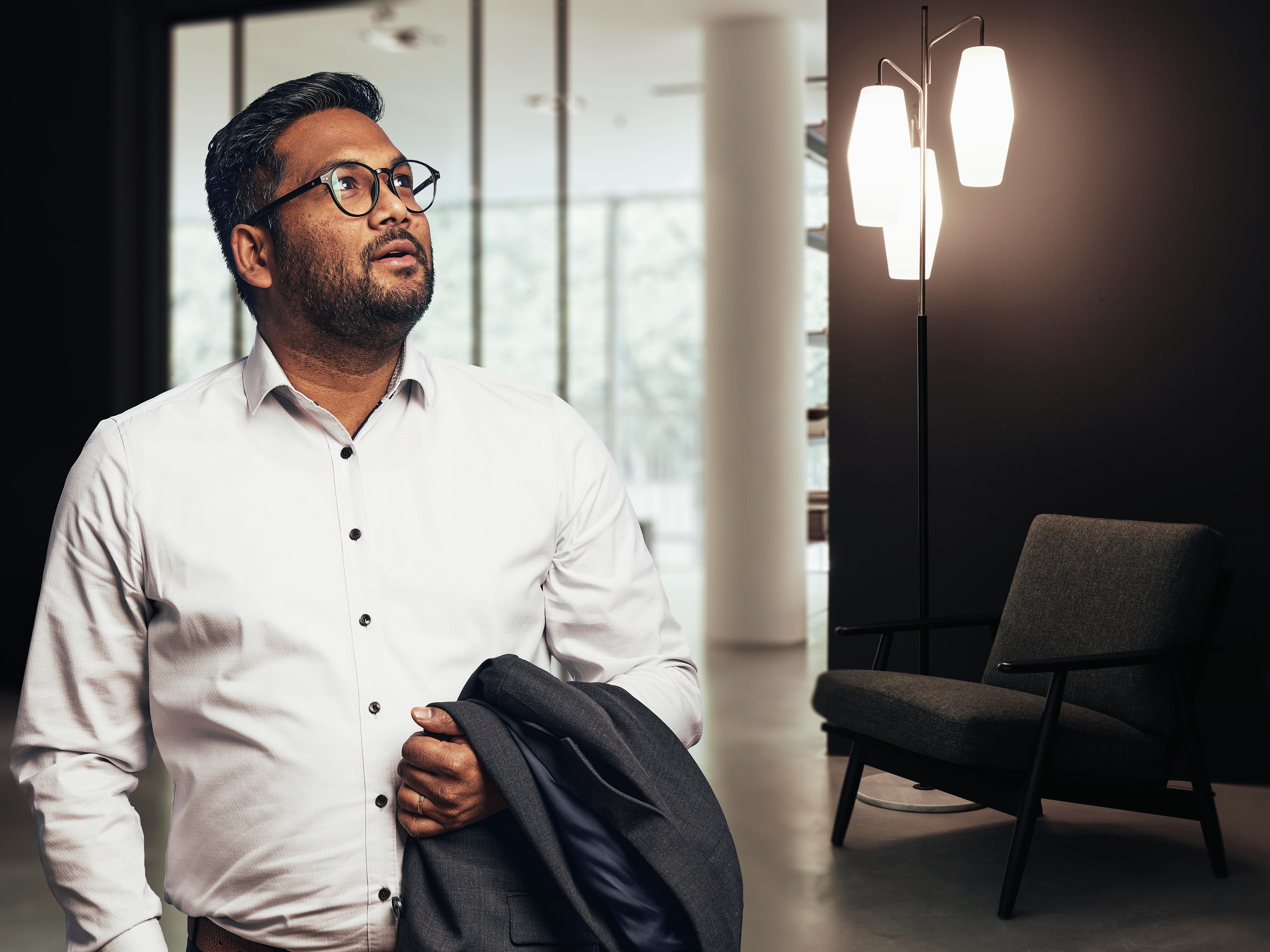 A man in a white shirt and glasses stands in an office, holding a jacket and looking upward.