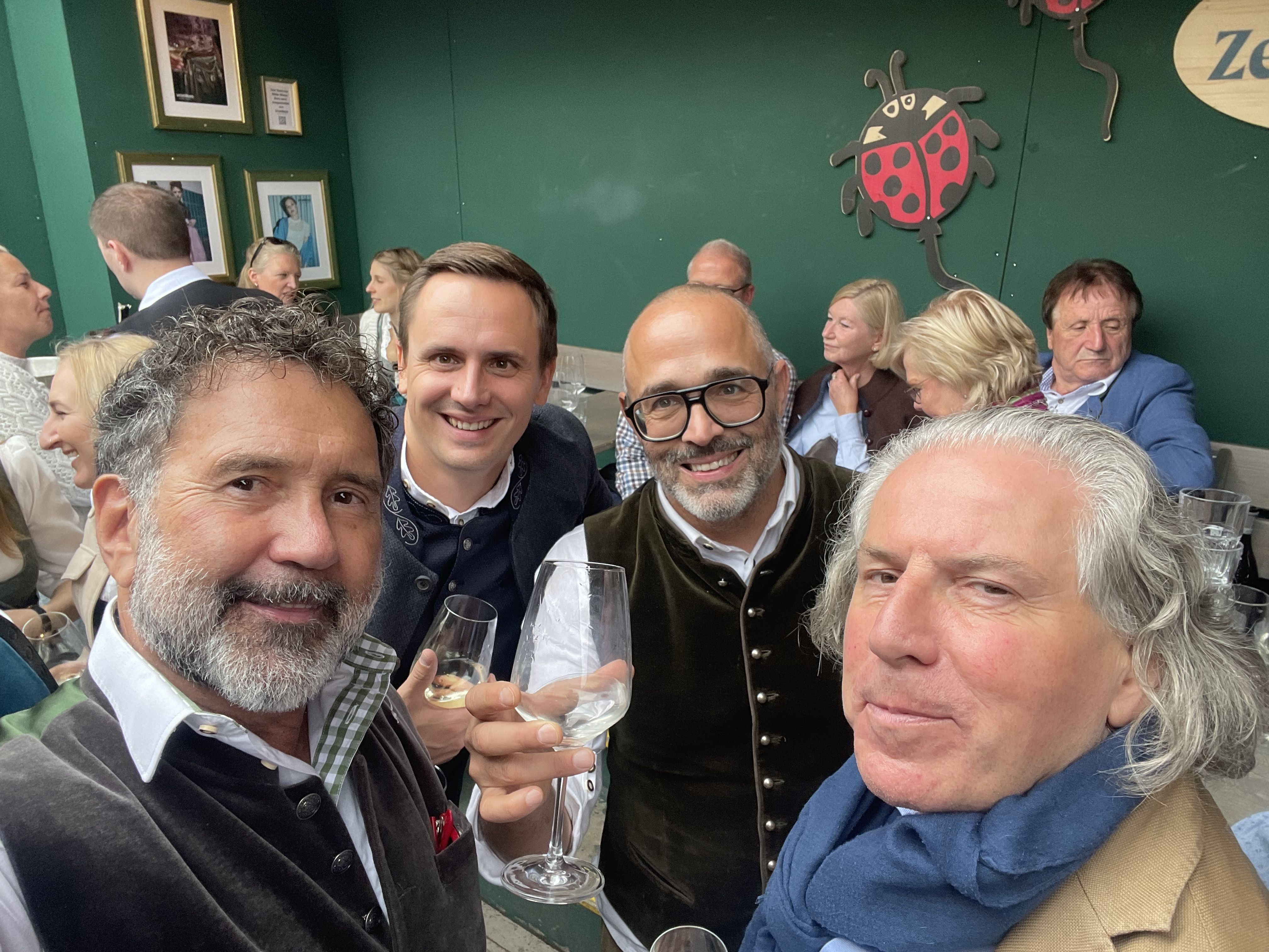 Quatro homens sorrindo e posando para uma foto em um restaurante, com um deles segurando uma taça de vinho.
