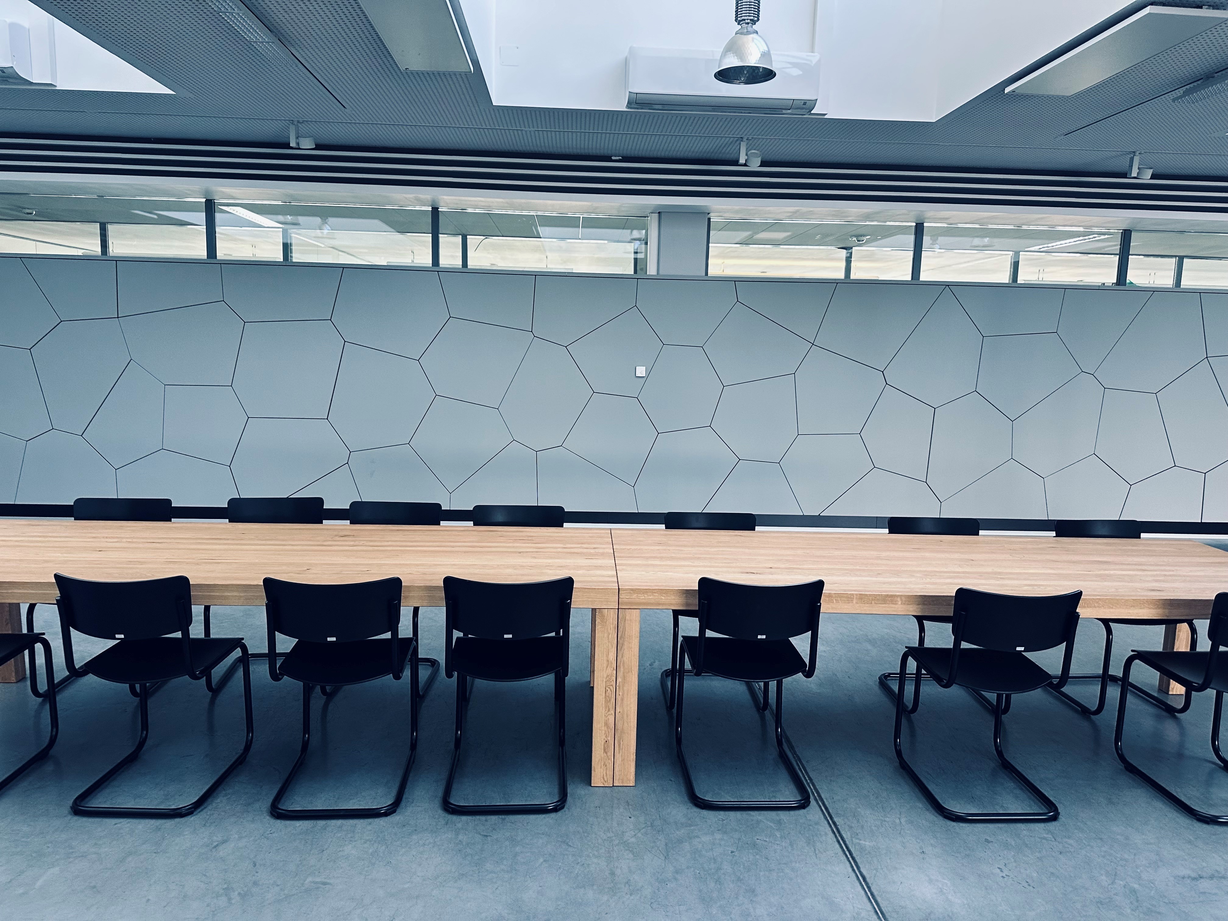 Long wooden table with black chairs arranged in a conference room with a hexagonal patterned wall and large windows.