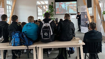 A group of students are sitting on benches in a classroom, watching a presentation on a projector screen.