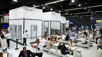 A large room with white walls and a white floor. There are several people sitting on benches and tables, and some are standing. There are also several people standing near the white walls. There are also several lights on the ceiling.